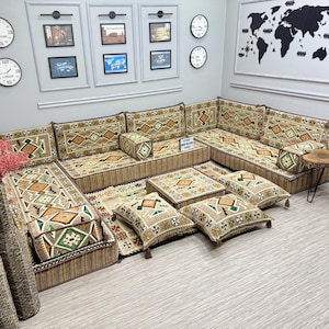 Puede incluir: Un gran sofá seccional beige con cojines estampados y una alfombra a juego. El sofá presenta diseños geométricos en tonos verdes, naranjas y marrones. Cojines decorativos y una pequeña mesa auxiliar de madera completan el conjunto.