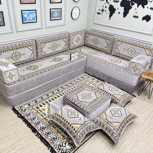 Op de afbeelding: Een grijze hoekbank met kussens met patronen en een bijpassend vloerkleed. De kussens en het vloerkleed hebben een geometrisch ontwerp in goud, zwart en oranje. De bank is in een hoek geplaatst, waardoor een gezellige zithoek ontstaat.