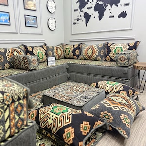 Op de afbeelding: Een grijze hoekbank met kussens met patronen en een bijpassende poef. De kussens hebben een geometrisch ontwerp in zwart, groen en oranje. Een kleine houten bijzettafel met zwarte metalen poten is ook zichtbaar.