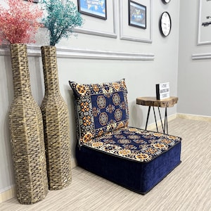 May include: Two tall, woven, bottle-shaped floor vases containing pink and teal dried flowers, positioned next to a low-profile, square floor cushion featuring a dark blue fabric with a repeating geometric and floral pattern in orange and white.