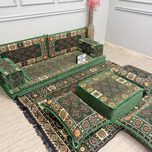 May include: A green and brown patterned floor seating set. The set includes a sofa, two armrests, a large square ottoman, and several throw pillows. The set is placed on a matching rug with a black fringe.