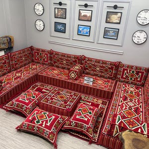 May include: A red and cream-coloured sectional sofa with matching floor cushions and pillows. The cushions and sofa feature geometric patterns. The room has framed artwork and clocks on the wall. The text on the table says "THE ANATOLIAN SOFA".