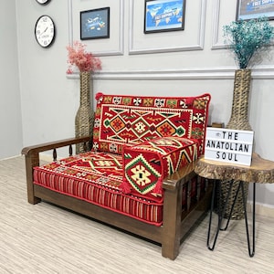 Puede incluir: Un sofá de madera con un cojín y respaldo estampados en rojo. El cojín presenta un diseño geométrico en rojo, verde y amarillo. El sofá tiene estructura y reposabrazos de madera. Una pequeña mesa auxiliar con el texto "THE ANATOLIAN SOUL".