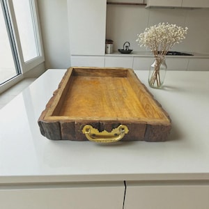 Puede incluir: Una bandeja rectangular de madera para servir con un interior de veta de madera natural y un exterior marrón oscuro, similar a la corteza. Cuenta con un asa dorada y está diseñada para servir o exhibir. La bandeja está sobre una superficie blanca.