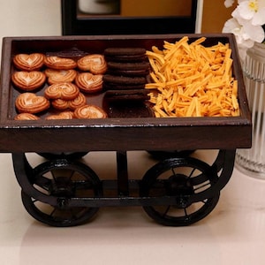 Puede incluir: Un carrito de madera en miniatura lleno de una variedad de golosinas. El carrito tiene un acabado marrón oscuro y ruedas negras. Las golosinas incluyen galletas en forma de corazón, galletas de chocolate y bocadillos dorados. Una pieza decorativa para servir aperitivos.