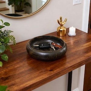 May include: A dark brown, circular wooden bowl with a distressed finish containing a set of metal keys with a brown leather keychain. The bowl has a smooth, rounded rim and a deep interior, providing a functional space for holding small items.