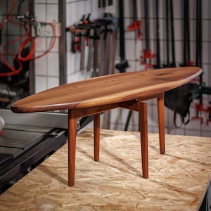 Pode incluir: Uma mesa de centro em estilo moderno de meados do século, com um tampo oval de madeira maciça e quatro pernas afiladas. A mesa é feita de madeira de tons quentes, provavelmente nogueira, e assenta numa superfície clara.