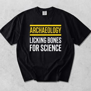 Peut inclure: Un t-shirt noir à manches courtes avec le texte jaune ARCHAEOLOGY centré entre deux lignes horizontales jaunes, et le texte blanc LICKING BONES FOR SCIENCE imprimé juste en dessous dans une police de caractères sans empattement.