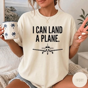 May include: An ivory short-sleeved t-shirt featuring the black text I CAN LAND A PLANE and a graphic of a small airplane. The shirt is paired with a white mug decorated with a repeating pattern of small blue palm trees.