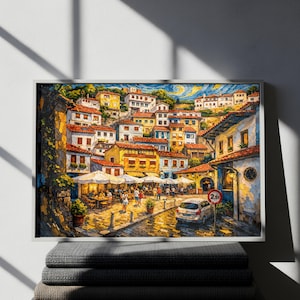Può includere: Stampa artistica incorniciata di una scena di un villaggio europeo. Il dipinto raffigura edifici colorati con tetti di tegole rosse, una strada acciottolata e persone che cenano in caffè all'aperto. Sono visibili anche un'auto e un segnale stradale.