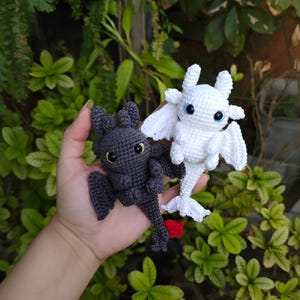 May include: Two crocheted dragon figures, one dark grey and one white, are shown against green foliage. The dark grey dragon has yellow eyes, while the white dragon has blue eyes. Both have wings and are handmade.