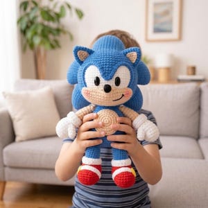 Könnte beinhalten: Ein handgefertigtes blaues Sonic the Hedgehog-Plüschtier aus Häkelarbeit. Das Spielzeug hat weiße Handschuhe, rot-weiße Schuhe und einen beigen Bauch und ein beiges Gesicht. Das Plüschtier wird von einer Person gehalten.