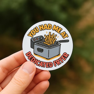 May include: A round sticker with the text "YOU HAD ME AT DEDICATED FRYER" in orange and red. The sticker features a cartoon illustration of a deep fryer with french fries. The fryer is gray with yellow accents.