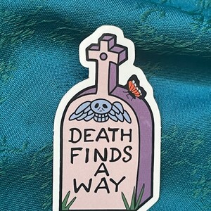 May include: A sticker featuring a tombstone design with the words "DEATH FINDS A WAY" in black lettering. The tombstone is light purple with a cross at the top and a skull with wings. A small butterfly rests on the side. Green grass is at the bottom.