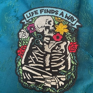 May include: Embroidered patch featuring a skeleton embracing, surrounded by colorful flowers and green leaves. The patch has the text "LIFE FINDS A WAY" on a light blue banner. The patch is on a teal fabric.