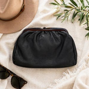 May include: Black leather clutch with a dark brown clasp. The clutch is on a cream-colored fabric, with a tan hat and tortoiseshell sunglasses in the background. A sprig of green leaves is also visible.