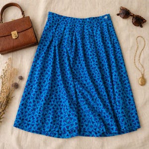 May include: A blue skirt with a geometric triangle pattern. The skirt is pleated and has a button closure. A brown purse, sunglasses, and a gold necklace are also in the image.
