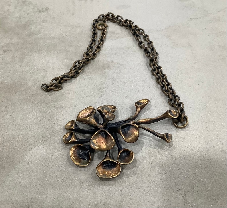 May include: A bronze necklace with a pendant shaped like a cluster of stylized flowers. The pendant is made of multiple interconnected pieces, each with a unique shape and texture.