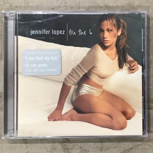 May include: A Jennifer Lopez On the 6 music CD in a clear plastic jewel case. The cover features a photograph of a person in a beige long-sleeve top and metallic silver shorts, with a sticker stating: features both hit singles If you had my love and No me ames (duet with Marc Anthony).