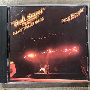 Puede incluir: Caja de CD del álbum "Nine Tonight!" de Bob Seger & The Silver Bullet Band. La portada presenta el nombre de la banda y el título del álbum en una fuente estilizada, con un escenario de concierto iluminado en rojo y naranja.