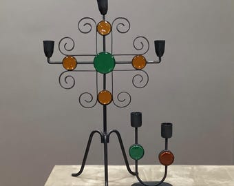 May include: Two black metal candle holders. One is a tall, cross-shaped design with orange and green glass accents and decorative swirls. The other is a smaller, curved holder with two candle cups and green and red glass accents.