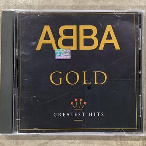 May include: A CD case for ABBA's "Gold: Greatest Hits" album. The cover is black with gold text and a crown design. The CD is housed in a clear plastic jewel case.