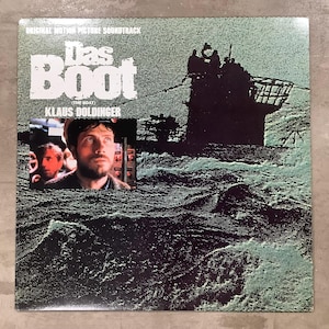 May include: A square vinyl record cover for the original motion picture soundtrack of Das Boot by Klaus Doldinger. The cover features a green and black halftone image of a submarine at sea, with a small inset photo of a bearded man in a cap.