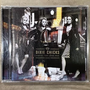 Op de afbeelding: CD-hoesje voor het Dixie Chicks-album "Taking the Long Way". De cover toont drie vrouwen in donkere jassen en jurken, bij een oldtimer. De albumtitel en de bandnaam zijn in het wit gedrukt.