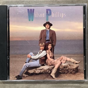 Puede incluir: Caja de CD con la portada del álbum de Wilson Phillips. La imagen muestra a tres mujeres posando sobre una formación rocosa con un fondo de puesta de sol. El nombre de la banda se muestra en texto turquesa y blanco en la parte superior.