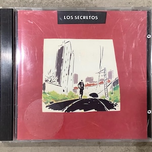 May include: A CD case with a red cover featuring a drawing of a person walking on a road with buildings in the background. The text "LOS SECRETOS" is at the top. The CD case is in a clear plastic wrap.
