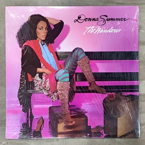 May include: A vinyl record album cover for Donna Summer's "The Wanderer" album. The cover features a woman in colorful 1980s attire, seated on a bench against a pink and black striped background. The album title and artist's name are in cursive script.