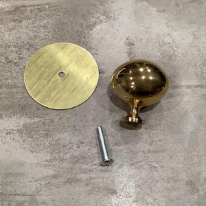 May include: A gold-toned round cabinet knob with a smooth, shiny finish. The knob has a small, round base and a screw for installation. A matching round base plate is also included.