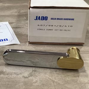 May include: A chrome and brass door handle with a rectangular body and a rounded end. The handle has a gold-coloured, ribbed accent on one end. The packaging box reads "JADO SOLID BRASS HARDWARE".