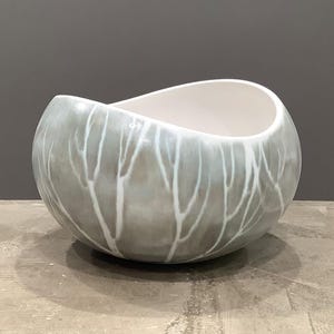 May include: A rounded ceramic bowl with an asymmetrical, undulating rim. The exterior features a soft, muted gray glaze with a white, branching, tree-like pattern that extends from the base toward the top. The interior of the bowl is smooth and white.