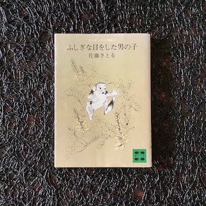 May include: A book cover with a light brown background and a black and white illustration of a person jumping. The title of the book is "ふしぎな日をした男の子供" and the subtitle is "佐藤さとる".
