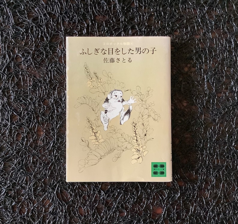 May include: A book cover with a light brown background and a black and white illustration of a person jumping. The title of the book is "ふしぎな日をした男の子供" and the subtitle is "佐藤さとる".