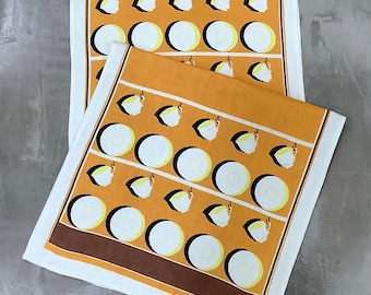 May include: Two white tea towels with a brown border. The towels have a repeating pattern of yellow and white circles and a stylized image of a teacup.