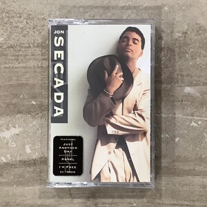 May include: A Jon Secada music cassette tape in a clear plastic case. The cover features a person in a light-colored suit holding a dark hat. Text on the cover reads JON SECADA and a black sticker lists the songs Just Another Day, Angel, and I'm Free.