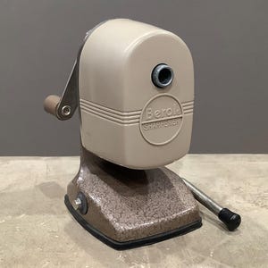 May include: A vintage Berol SHARPENER pencil sharpener in beige and brown. The sharpener has a hand crank and a textured base. The text "Berol SHARPENER" is printed on the front of the sharpener.