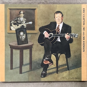 May include: Album cover art for "Eric Clapton | Me and Mr Johnson." The artwork features a seated figure playing a guitar, with two framed portraits in the background. The cover has a light tan border and the title is printed vertically.