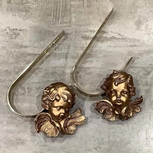 May include: Two gold cherub figurines with wings, one with a crown, on a white background. The cherubs are attached to metal hooks.