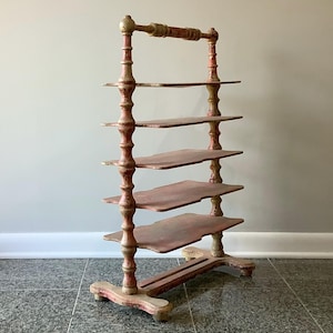 May include: An antique, multi-tiered shelf with a distressed, painted finish. The shelf has six levels, supported by ornate, turned posts. The color scheme includes red, cream, and gold accents, giving it a vintage aesthetic. The shelf is free-standing.