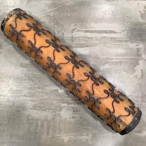 May include: A wooden rolling pin with a decorative metal pattern. The rolling pin is cylindrical with a light brown wood finish and dark metal accents. The metal pattern features a repeating floral design. The ends of the rolling pin are black.
