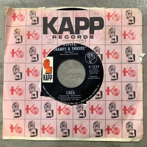 May include: Vintage 45 RPM vinyl record by Cher, titled "Gypsys, Tramps & Thieves." The record label is black with white text, and the sleeve is pink with a repeating pattern of "K" logos and illustrations.