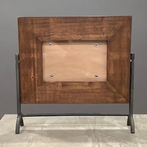 May include: A rectangular wooden picture frame with a dark brown finish. The frame is supported by a dark gray metal stand. The frame has a light brown backing and is held together with screws. The design is simple and modern.