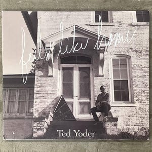 Può includere: Una copertina quadrata di un album musicale con una fotografia in bianco e nero di una casa in mattoni. Una persona è seduta sui gradini accanto a uno strumento triangolare. Il testo riporta feels like home e Ted Yoder in bianco.