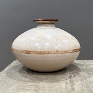 Puede incluir: Un jarrón de cerámica color crema con cuerpo redondeado y cuello estrecho. El jarrón presenta una banda marrón en el centro y un borde marrón. El jarrón está sobre una superficie gris.