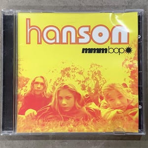 May include: A CD case featuring the band Hanson. The album cover is yellow with the band's name in orange and the song title "mmmbop". The image shows three people lying in grass.