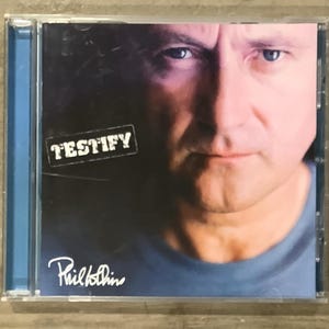 May include: A music CD in a clear plastic jewel case featuring the album Testify by Phil Collins. The cover art displays a close-up portrait of a man with blue eyes, wearing a blue shirt, with the word TESTIFY printed in white on a dark background.