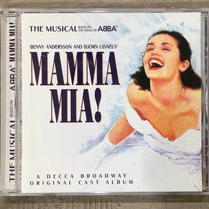 May include: A CD case for the musical "Mamma Mia!" based on the songs of ABBA. The cover features the title in large, dark blue letters, with a smiling woman in a white dress. The text includes "Benny Andersson and Björn Ulvaeus'" and "A Decca Broadway Original Cast Album."